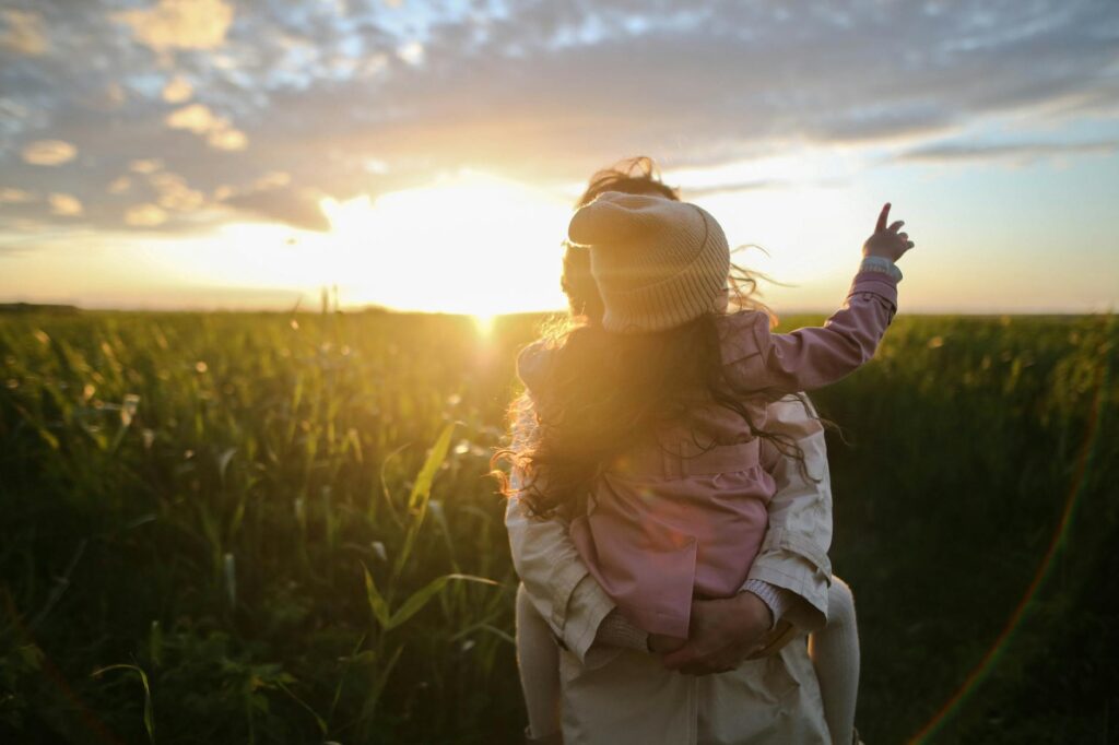 A beautiful sunset behind a mom carrying a young girl as the girl points to something in the distance.